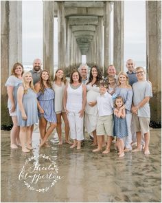 a large family poses for a photo under the pier in front of the ocean on a cloudy day