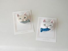 two white cats with blue bow ties on their heads, one in a card and the other in a fake cat's head