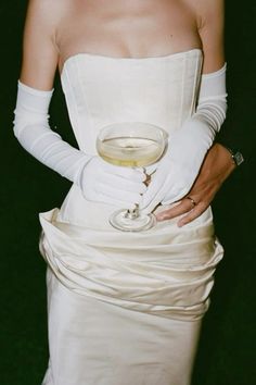 a woman in a white dress holding a glass of wine and wearing gloves on her arm