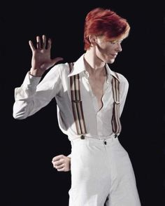 a man with red hair wearing white pants and suspenders, making the peace sign