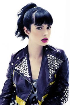 a woman with black hair wearing a purple leather jacket and yellow accents is posing for the camera