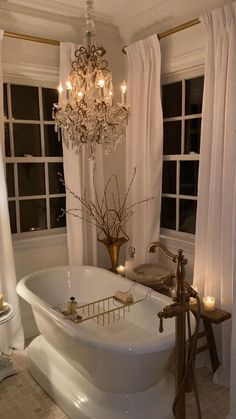 a white bath tub sitting under a chandelier in a bathroom next to two windows