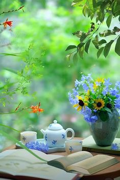 an open book on a table next to a vase filled with flowers and tea cups