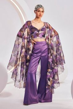 Purple long cape with intertwined floral bloom print. Paired with an inner padded blouse with all over floral bloom print. Paired with a trouser.
Components: 3
Pattern: Print
Type Of Work: Floral Bloom
Neckline: V Neck
Sleeve Type: Sleeveless
Fabric: Blouse: Printed Chiffon, Pant:Heavy Satin, Cape:Printed Organza
Color: Purple
Other Details: 
Attached lining
Blouse:
Padded
Crystal stone lace hem
Occasion: Sangeet - Aza Fashions Chic Floral Print Festive Sets, Chic Floral Print Sets For Festive Occasions, Chic Festive Sets With Floral Print, Silk Floral Print Party Sets, Summer Party Palazzo Set With Cape Sleeves, Elegant Summer Cape Set, Elegant Georgette Palazzo Set With Floral Print, Summer Evening Sets With Cape Sleeves, Spring Silk Set With Kimono Sleeves