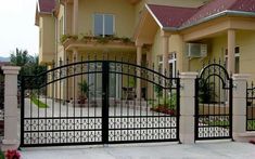 an iron gate in front of a house with flowers on the ground and bushes around it