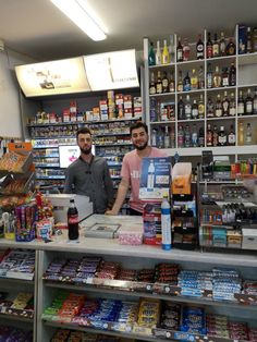 two men standing behind a counter in a store filled with drinks and condiments