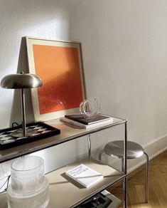 a glass table topped with a lamp next to a white chair and a painting on the wall