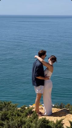 a man and woman standing next to each other on top of a cliff near the ocean