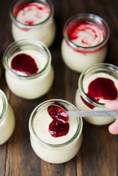 small jars filled with white and red jello on top of a wooden table next to a hand holding a spoon