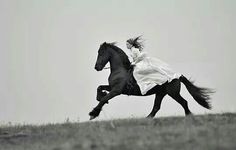 a woman riding on the back of a black horse in a field with tall grass