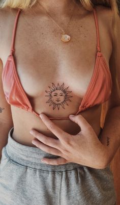 a woman with a sun tattoo on her chest wearing a pink bra and grey sweatpants