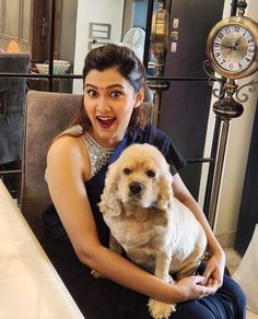 a woman holding a dog in her lap while sitting at a table with a clock behind her