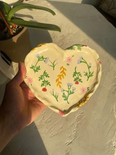 a hand holding a heart shaped dish with flowers painted on the front and sides, next to a potted plant