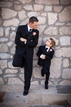 two young boys dressed in suits and bow ties, standing next to each other against a stone wall