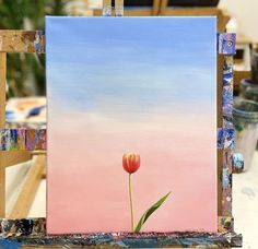 an easel with a painting on it and a flower in the foreground, against a pink sky