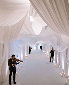 two men in tuxedos are playing violin on a white wedding aisle with candles