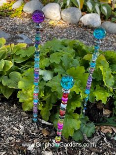 three colorful glass candlesticks sitting on top of a garden bed next to green plants