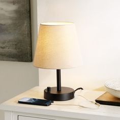 a table lamp sitting on top of a white dresser next to a phone and bowl