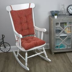 a white rocking chair with an orange cushion next to a gray cabinet and clock on the wall