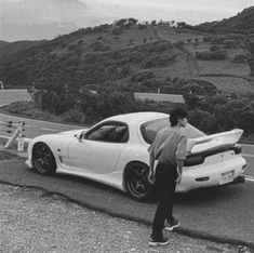 a man standing next to a white sports car on the side of a mountain road