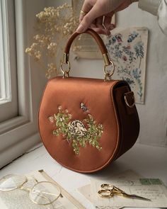 Embrace the essence of spring Sleepy Fawn Embroidered Saddle Bag. Crafted from luxurious brown satin, this bag features a rounded, saddle-shaped silhouette adorned with detailed embroidery depicting a sleeping fawn nestled amongst the wildflowers of spring—an embodiment of tranquillity and the gentle beauty of nature's renewal. Designed for both beauty and functionality, the bag comes complete with a top handle for easy carrying and an adjustable cross-body strap for hands-free convenience. The Bride Glam, Fable England, Luxury Christmas Gifts, Spring Wildflowers, Detailed Embroidery, Retro Bags, Hand Painted Jewelry, August Birthstone Jewelry, Brown Satin