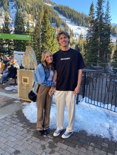 a man and woman standing next to each other in front of snow covered mountains with trees