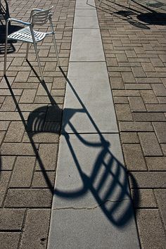 two chairs and a table on a bricked sidewalk with the shadows of them casting a long shadow