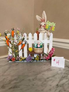 an easter decoration with flowers and eggs on a table in front of a white picket fence