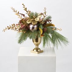 a gold vase filled with flowers and greenery on top of a white block in front of a white wall