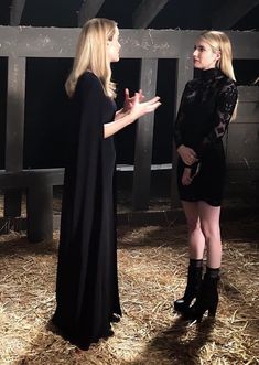 two women standing in hay talking to each other with one woman wearing a black cape