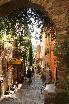 people walking down an alley way with shops on either side and trees growing over the walkway