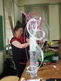 a woman standing next to a tall glass vase filled with water and streamers on top of a wooden table