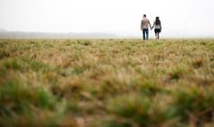 two people walking in the grass holding hands and looking at something on a foggy day