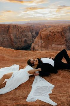 a man and woman laying on top of a rock in front of the desert at sunset