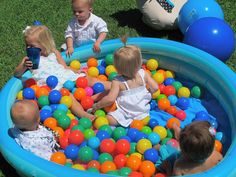 three children playing in an inflatable pool filled with balls and drinking water while another child looks on