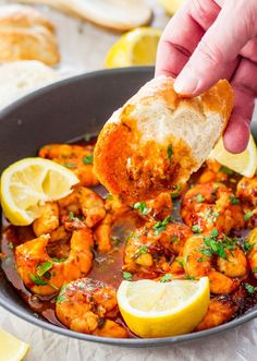 a hand dipping a piece of bread into a skillet filled with shrimp and lemons