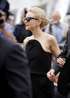 a woman in a black dress and sunglasses walking next to a man