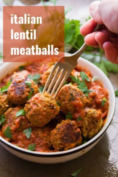 a hand holding a fork over a bowl of meatballs with sauce and parsley