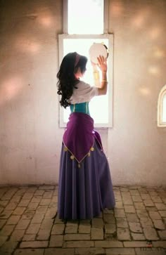 a woman in a long purple dress looking out a window at the sun coming through