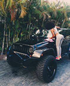 a woman standing on the hood of a black jeep in front of trees and palm trees