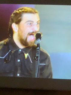 a bearded man singing into a microphone in front of a screen with the image of him on it