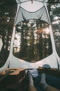 a person laying in a tent with their feet propped up on the ground and looking out into the woods