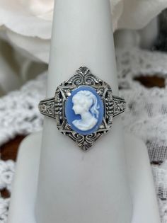 Blue Lady Cameo RingCleopatra Design#D209 Discover the classic beauty of Egyptian Revival with this gorgeous Cameo Ring. Crafted of sterling silver, this exquisite Art Deco reproduction resembles a scene from another time in history. The simulated oval cameo is 10mm x 8mm and set into an 18mm North to South band. The lovely ivory silhouette of a graceful lady is set on a soft blue background. Graced by intricate filigree and swirl patterns, the geometric border will be sure to have heads turning Blue Cameo Jewelry For Wedding, Victorian Blue Cameo Jewelry, Collectible Blue Rings, Blue Cameo Jewelry For Formal Occasions, Formal Blue Cameo Jewelry, Graceful Lady, London Blue Topaz Earrings, Rainbow Opal, Filigree Necklaces