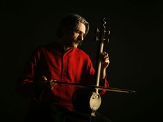 a man in red shirt holding an instrument and looking at the camera with dark background