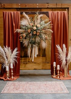 an arrangement of flowers and feathers on display in front of a red curtain with candles