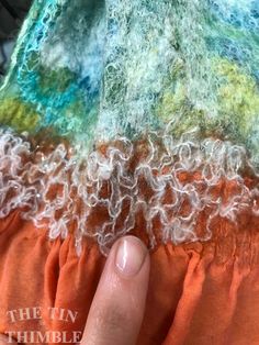 the finger is on top of an orange and green piece of fabric with white thread