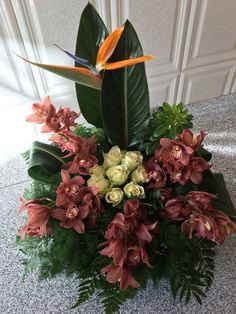 an arrangement of flowers and greenery in a vase on a counter top with a bird of paradise