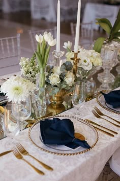 the table is set with white flowers and gold place settings, blue napkins and silverware