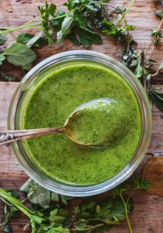 a spoon in a jar filled with green liquid surrounded by fresh herbs and greenery