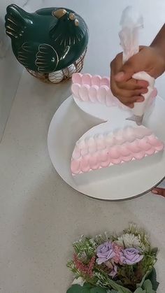 someone is decorating a cake with pink icing on a white plate next to flowers